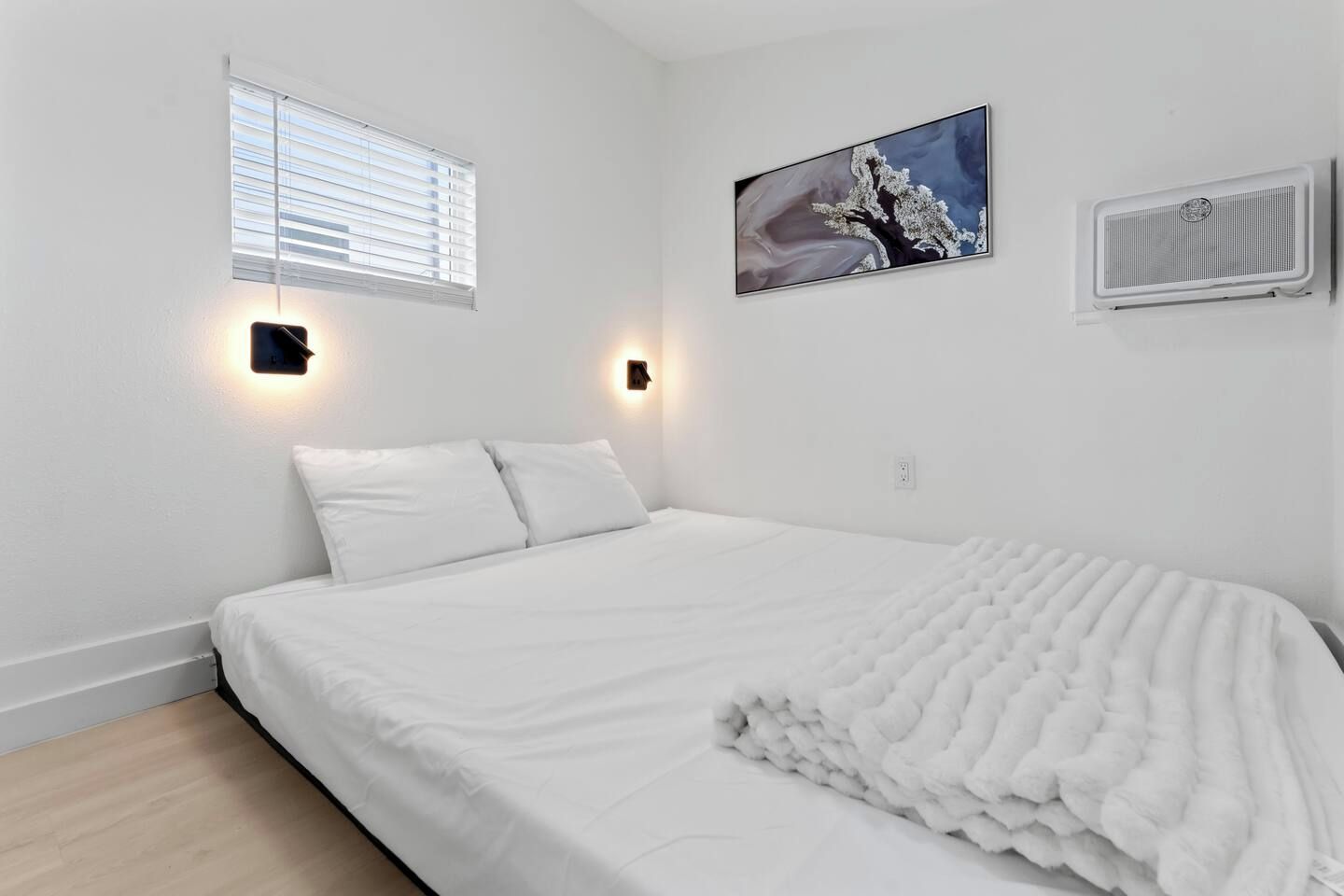 Een slaapkamer met een bed, twee wandlampen, een raam met zonwering en een airconditioner.