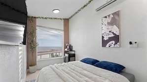A bedroom with a large bed, a window with a view, a wall-mounted air conditioner, and a painting. - Waterfront Tiny Suite | Cal King + Bay Views|Kemah (Bacliff)