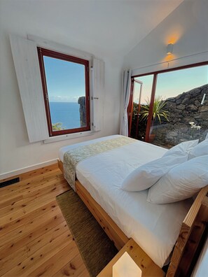 A bedroom with a wooden floor, a bed with white bedding, a window with curtains, and a view of the sea and rocks. - Mint and Honey - Azorean architecture with modern elegance and a 180° ocean view (Lajes do Pico)