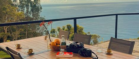 A wooden table with chairs, a table umbrella, and various items including a book, a camera, and a bowl of fruit.