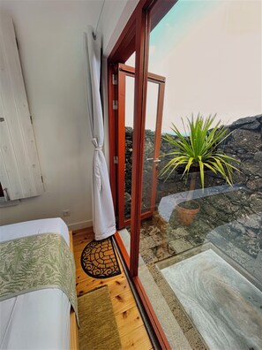 Quarto com cama, cortina branca, piso de madeira e porta de vidro que dá para área externa com planta.