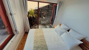 A bedroom with a bed, white pillows, a wooden headboard, a window, and a view of a stone wall and plant outside. - Mint and Honey - Azorean architecture with modern elegance and a 180° ocean view (Lajes do Pico)