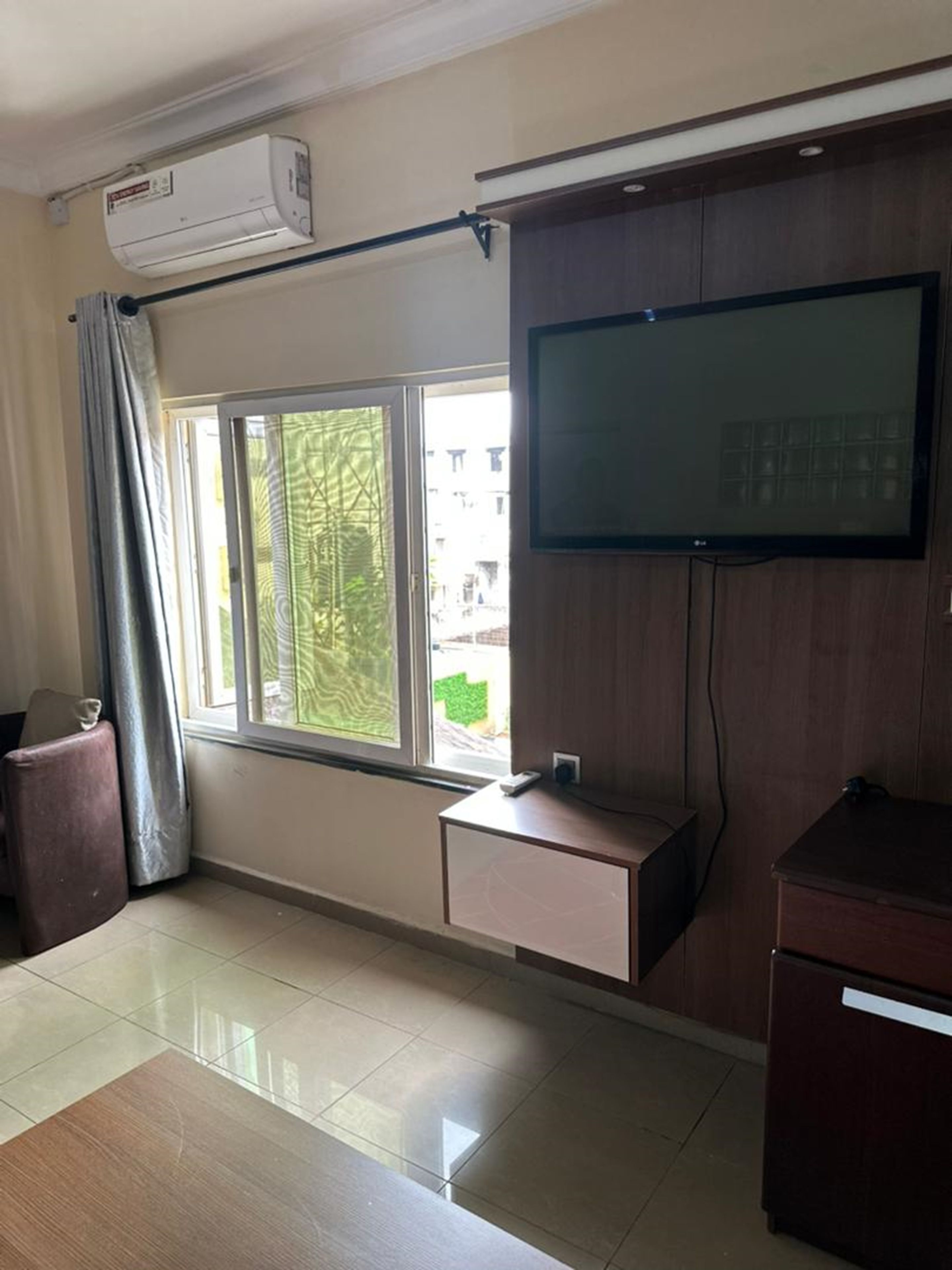 A room with a large window, a flat-screen TV mounted on the wall, a small cabinet, and a ceiling-mounted air conditioning unit.