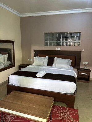 A hotel room with a large bed, a bedside table, a nightstand, a mirror, and a window with blinds. - Signatious hotel and Suite Abuja (Abuja)