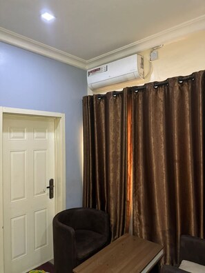 A room with a white door, a brown curtain, a brown sofa, and a wooden table. - Signatious hotel and Suite Abuja (Abuja)
