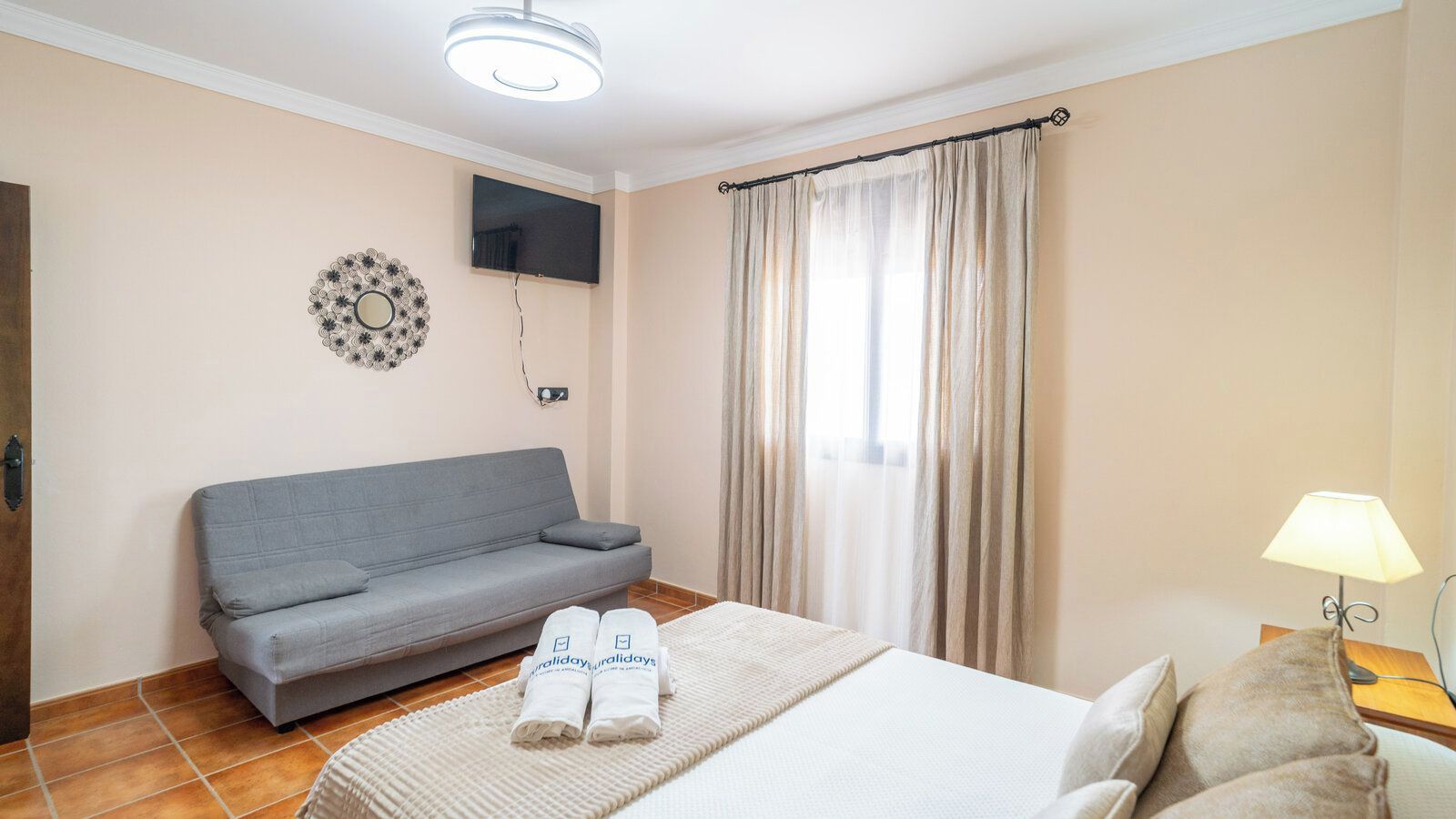 Une chambre d’hôtel avec un lit, un canapé gris, un téléviseur fixé au mur et une fenêtre avec des rideaux.