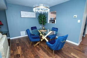 Ein Essbereich mit einem Glastisch und blauen Samtstühlen, einer Chandelierleuchtre und einer durch einen Bogen sichtbaren Küche.