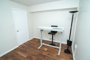 Quarto com porta branca, escrivaninha branca com computador, luminária de chão preta e piso de madeira.