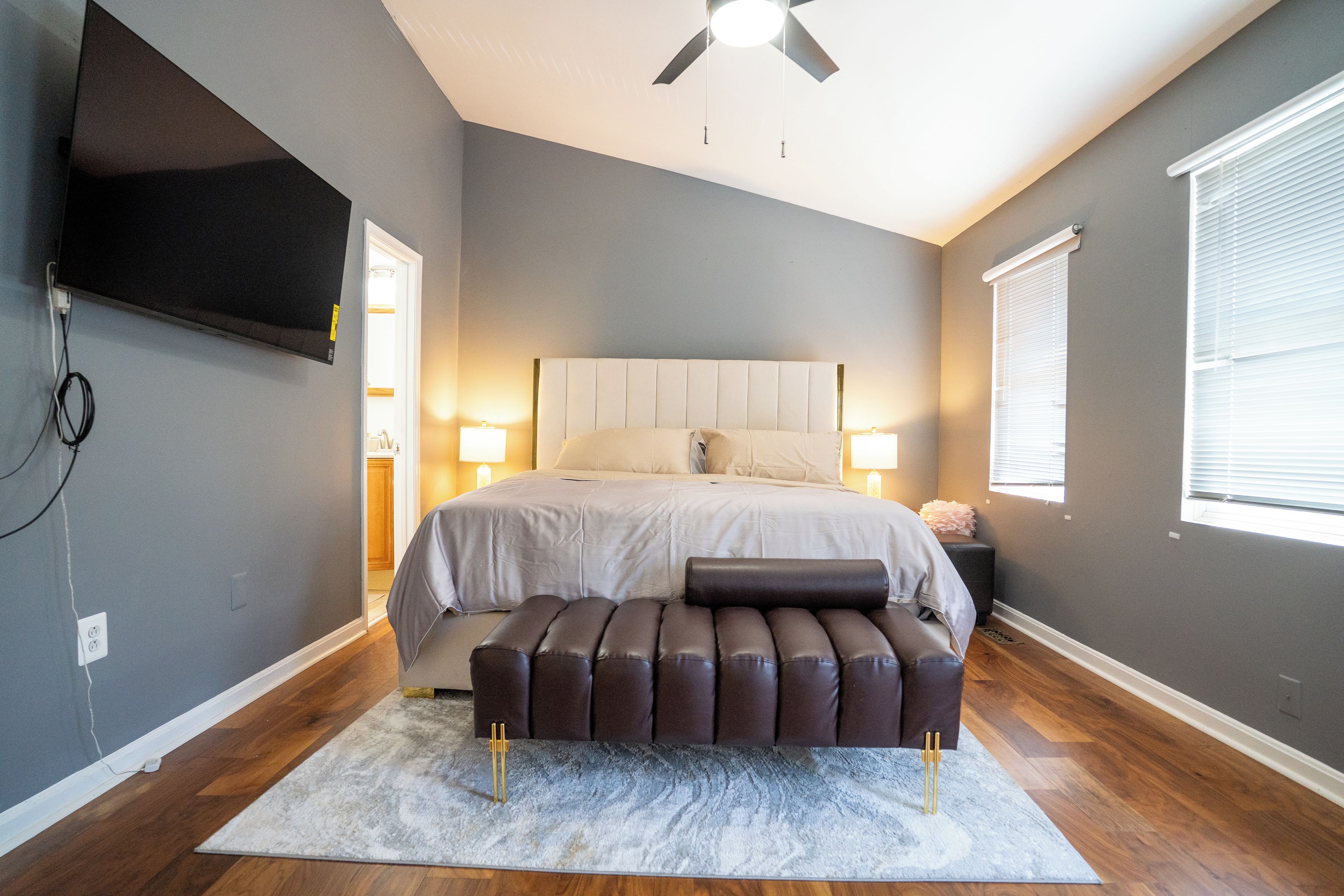 A bedroom with a bed, bedside lamps, a ceiling fan, a TV mounted on the wall, and a window with blinds.