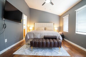 Ein Schlafzimmer mit einem Bett, Nachttischlampen, einem Deckenventilator, einem an der Wand montierten Fernseher und einem Fenster mit Jalousien.