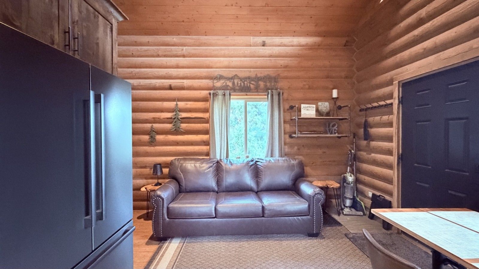 Ein Interieur einer Blockhütte mit einem Ledersofa, Holzschränken, einem Tisch und einem Fenster mit Vorhängen.