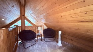 Quarto de cabana de madeira com teto inclinado, duas cadeiras pretas, uma mesa pequena e um aquecedor branco.