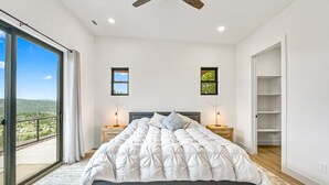 A bedroom with a large bed, two bedside tables, a ceiling fan, and a view of the outdoors. - Bryce Gateway Retreat — 2BR Cabin with Hot Tub & Mountain Views, Between Bryce & Zion (Alton)