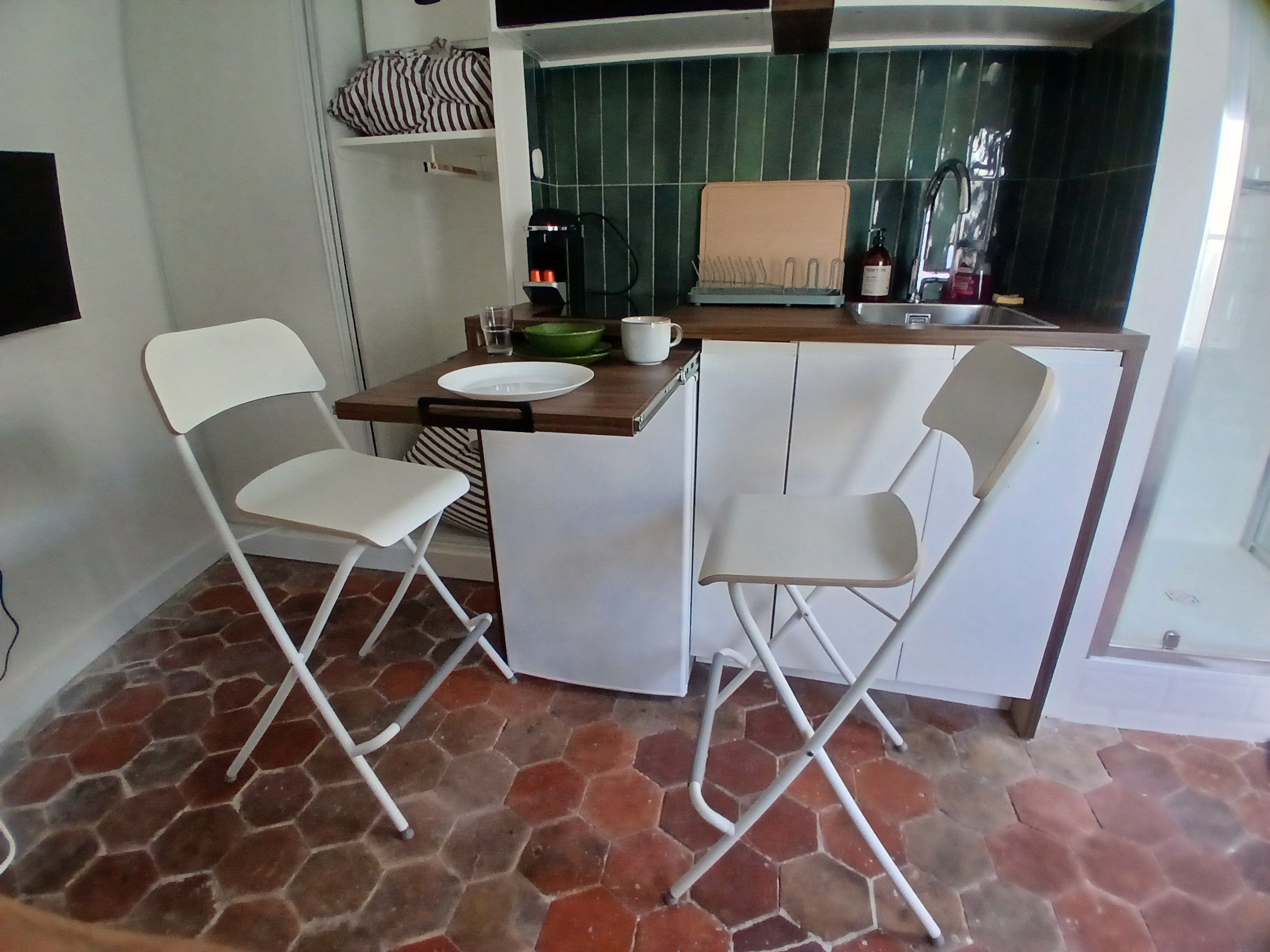 A compact kitchen with a dining area, including a white countertop, two white chairs, and a green tiled backsplash.