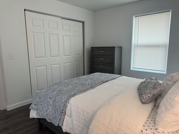 A bedroom with a bed, a dresser, a window with blinds, and a door.