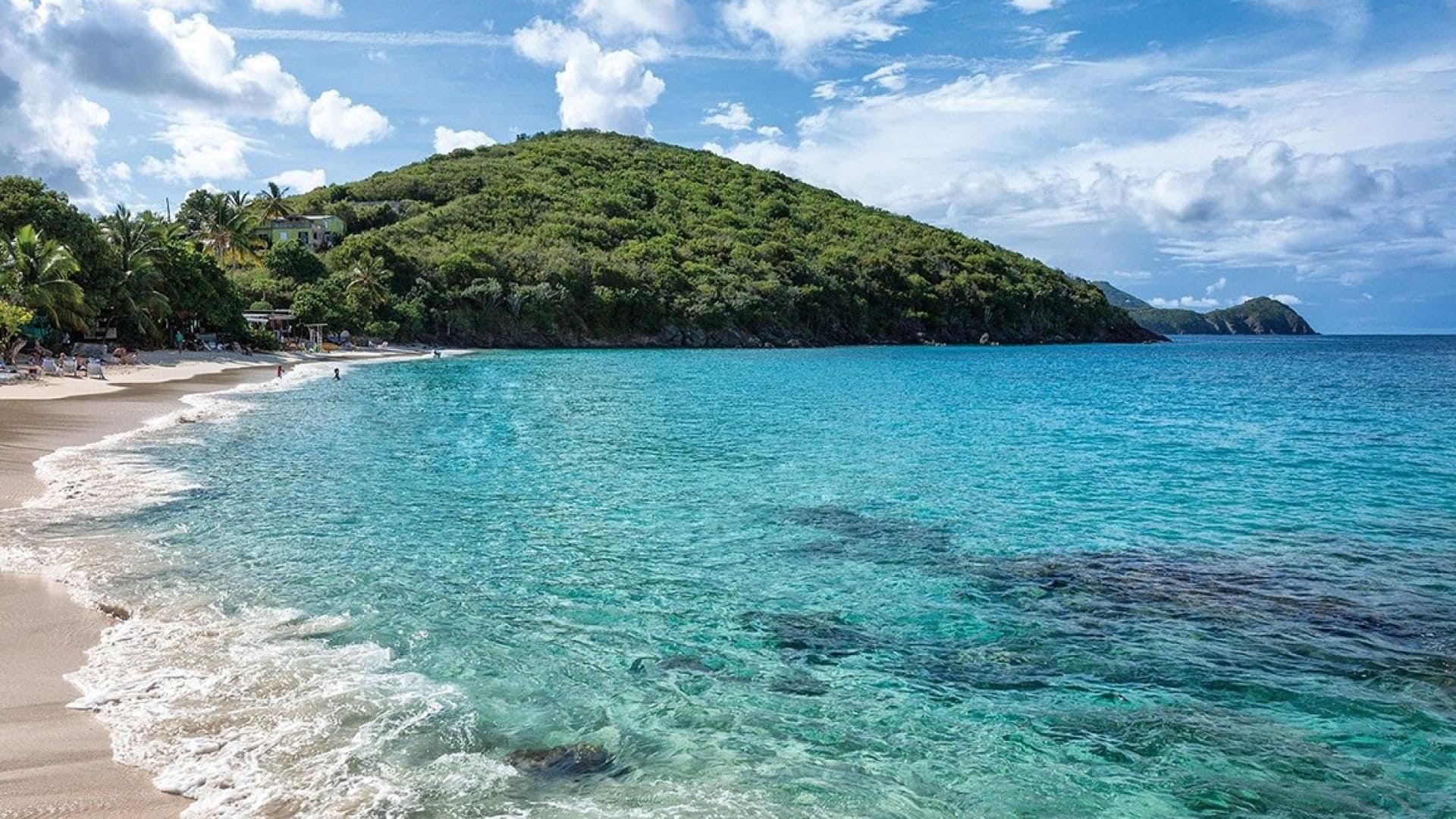Una platja amb aigua turquesa cristal·lina, sorra daurada i un turó cobert de vegetació verda abundant.