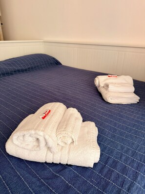 A bed with a blue quilted bedspread and two neatly folded white towels with a red logo. - WALK37 Syracuse - Between the Greek Theater and Ortigia (Siracusa)