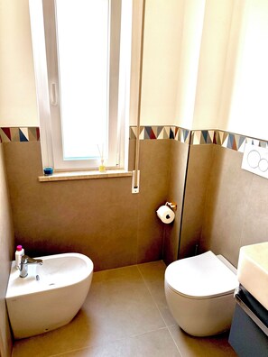 A bathroom with a toilet, bidet, and sink, a window with curtains, and a towel rack. - WALK37 Syracuse - Between the Greek Theater and Ortigia (Siracusa)