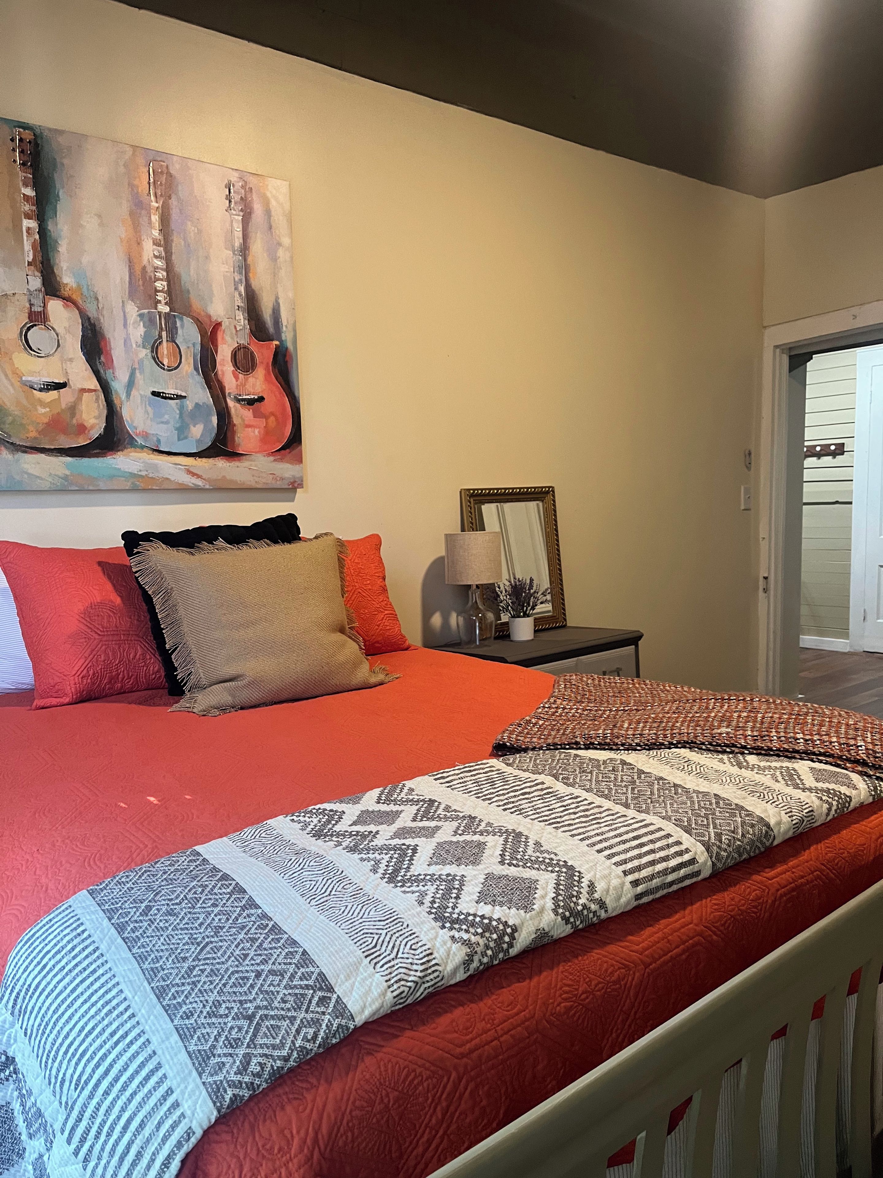 A bedroom with a large bed, a painting of guitars, a mirror, and a nightstand.