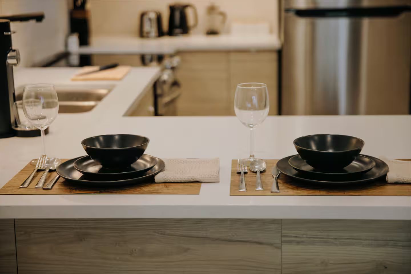 A modern kitchen with two dining sets on a white countertop, each with a black bowl, plate, and cutlery.