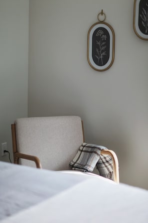 Ein Zimmer mit einem Bett, einem Sessel mit Kissen in Karomuster und zwei gerahmten Pflanzenbildern an der Wand.