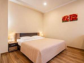 A neatly made bed with a headboard, a bedside table with a lamp, and two wall-mounted paintings. - Standart Double Room (Rīga)