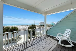 A balcony with a view of the beach and ocean, a white rocking chair, and a teal-colored wall. - Luxury Beach House 50 Steps To Beach / Heated Pool (Gulf Shores)