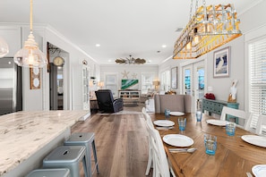 A spacious dining area with a wooden table set for six, a bar area with stools, and a modern kitchen with a marble countertop. - Luxury Beach House 50 Steps To Beach / Heated Pool (Gulf Shores)