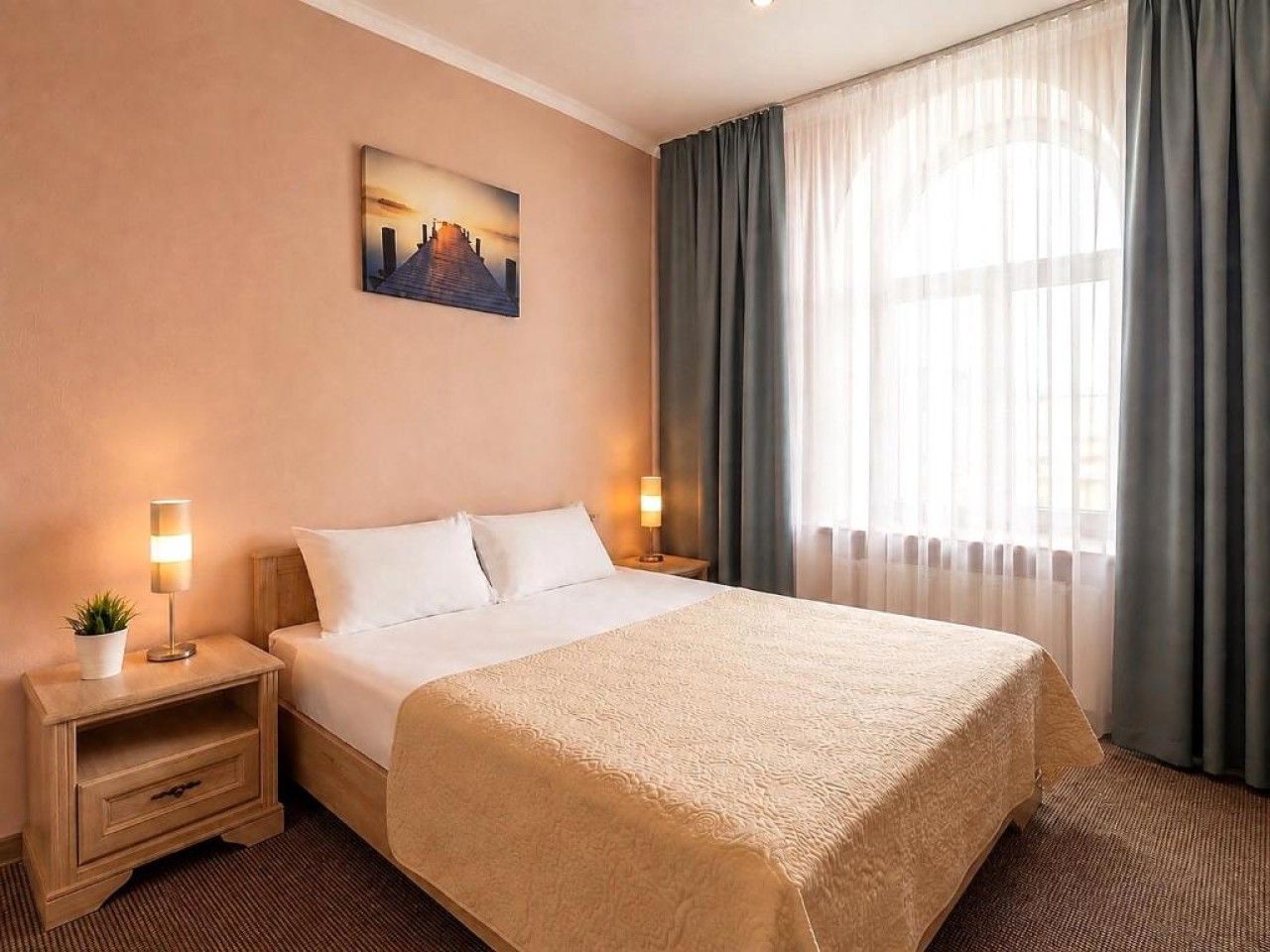 A hotel room with a large bed, a nightstand with a lamp, a painting on the wall, and a window with curtains.