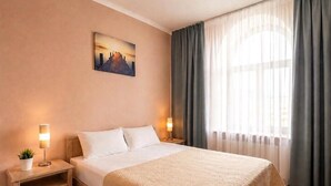 Una habitación de hotel con una cama grande, una mesita de noche con lámpara, un cuadro en la pared y una ventana con cortinas.