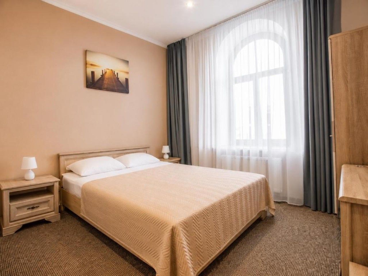 Ein Hotelzimmer mit einem Bett, einem Nachttisch, einer Lampe, einem Bild an der Wand und einem Fenster mit Vorhängen.