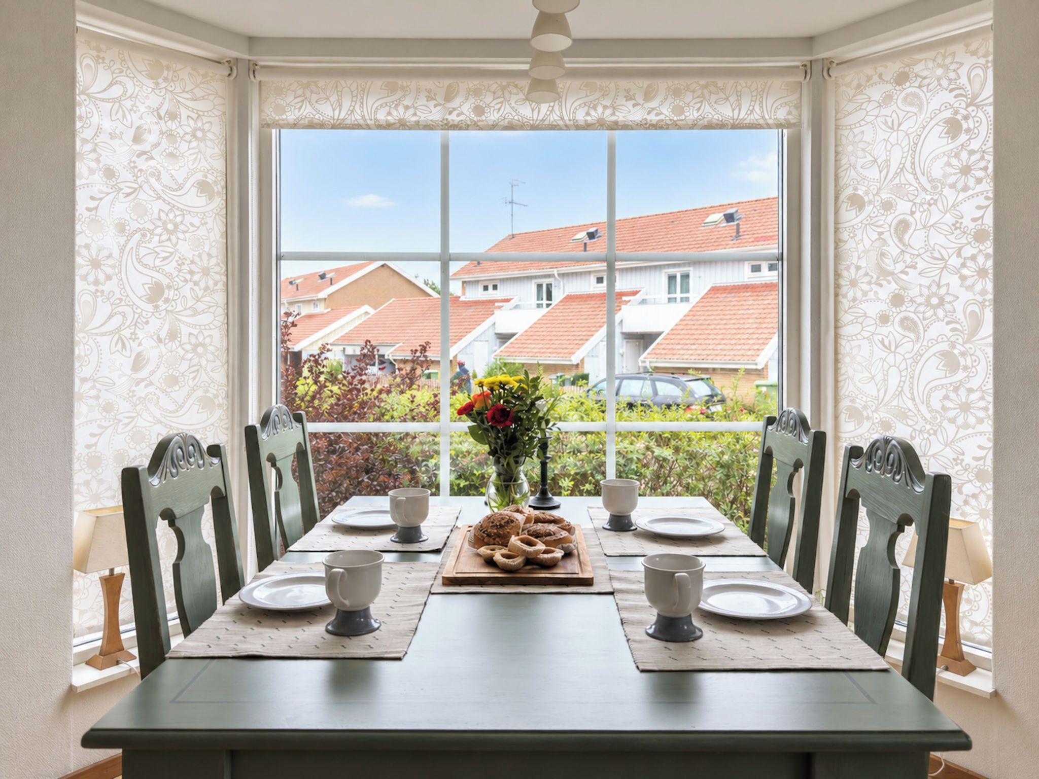 A dining table set for four with chairs, a vase with flowers, and a tray of pastries.