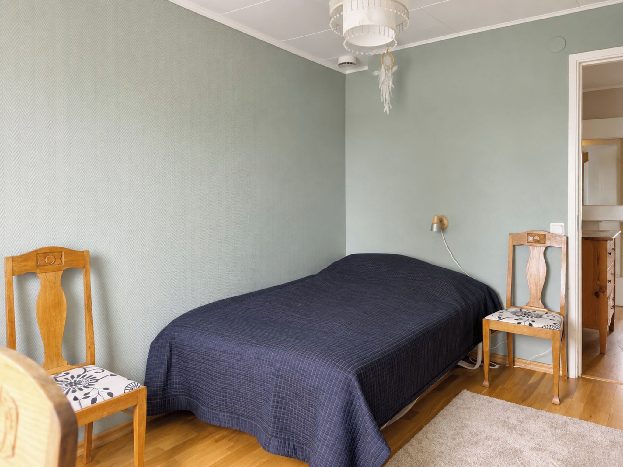 Ein Zimmer mit einem Bett, zwei Holzstühlen, einer weißen Deckenleuchte und einer Tür zu einem weiteren Raum.