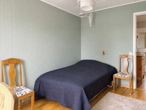 Una habitación con una cama, dos sillas de madera, una lámpara de techo blanca y una puerta que da a otra habitación.