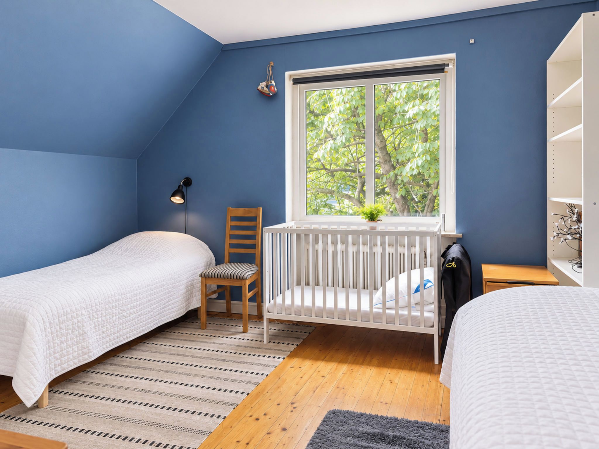 Una habitación con dos camas individuales, una cuna, una ventana con vistas a árboles, una silla y una mesita.