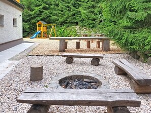 A fire pit area with benches, a children's play structure, and a playground in the background. - Pod Smrkem by Interhome (Janov nad Nisou)