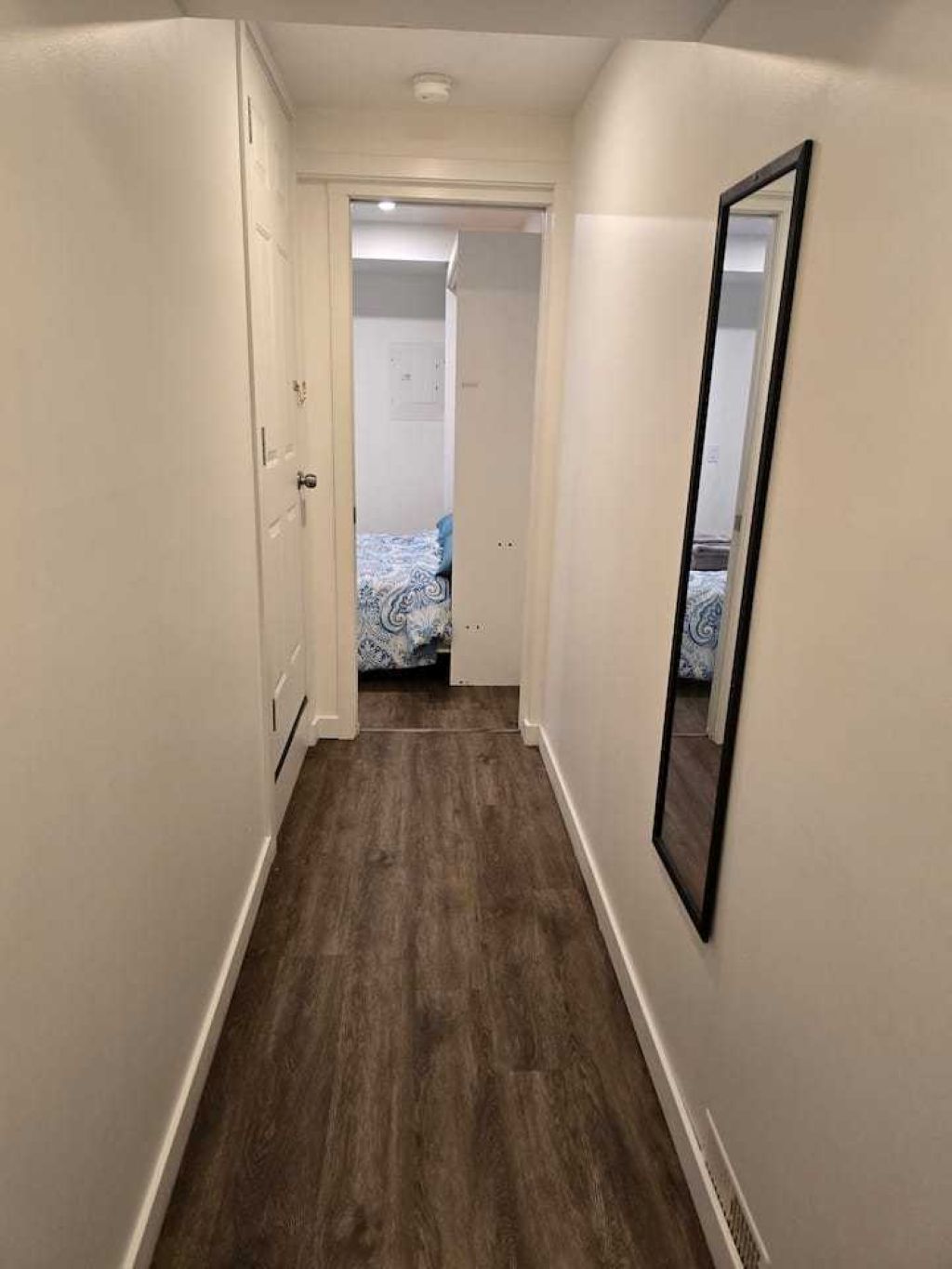 A narrow hallway with wooden flooring, white walls, and a door leading to a bedroom with a bed.