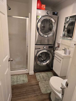 A compact laundry room with two stacked front-loading washing machines, a small sink, and a toilet. - Private Studio/bachelor suite (Victoria)