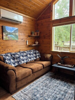 A wooden cabin interior with a brown couch, a blue patterned throw, a small table with a vase, and a painting of a lake.