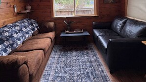 A cozy cabin interior with a ceiling fan, two windows, a brown couch, a black leather sofa, and a patterned rug. - Little Strawberry Suites, Tiny Home Downtown Prairie City (Prairie City)