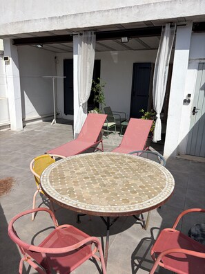 Un patio exterior con una mesa redonda y cuatro sillas, una planta en maceta y una puerta que da al interior.