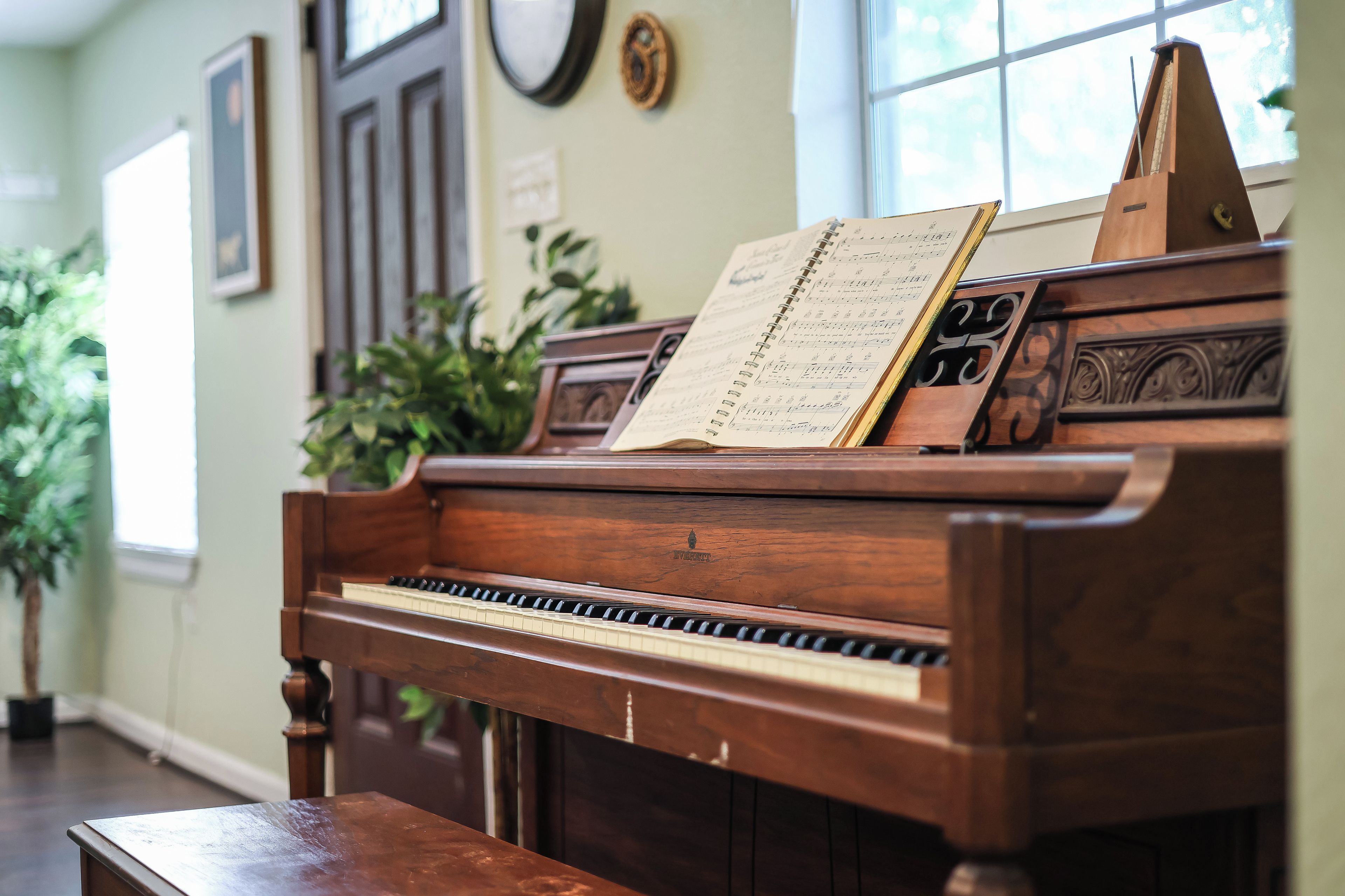 Um piano de cauda com partituras em uma sala com piso de madeira, uma planta em vaso e uma janela.