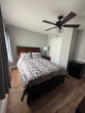 A bedroom with a bed, a ceiling fan, a nightstand, a lamp, a wardrobe, and a refrigerator.