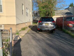Een woonstraat met een geparkeerde auto en een rode bestelwagen, een hek van gaas en een huis met een beige gevel.