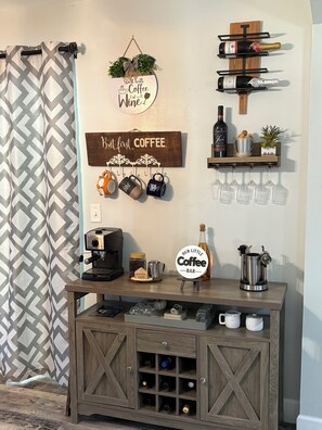 A coffee bar with a coffee machine, mugs, wine glasses, and a sign that reads 'Our Little Coffee Bar.'