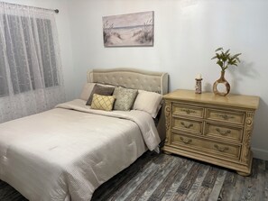 Un dormitorio con una cama, una mesita de noche, un cuadro y una ventana con cortinas traslúcidas.