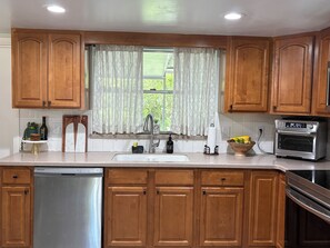 A kitchen with wooden cabinets, a stainless steel dishwasher, a built-in oven, and a window with patterned curtains.