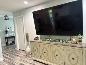 Una sala de estar con un televisor de pantalla plana grande montado en la pared, un mueble de madera con objetos decorativos y un pasillo que conduce a otra habitación.