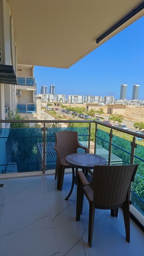 Parvekkeella on kaksi rottingista tuolia ja pieni pöytä, ja sieltä avautuu näkymä modernien rakennusten täyttämälle kaupunkimaisemalle ja kirkkaalle taivaalle.