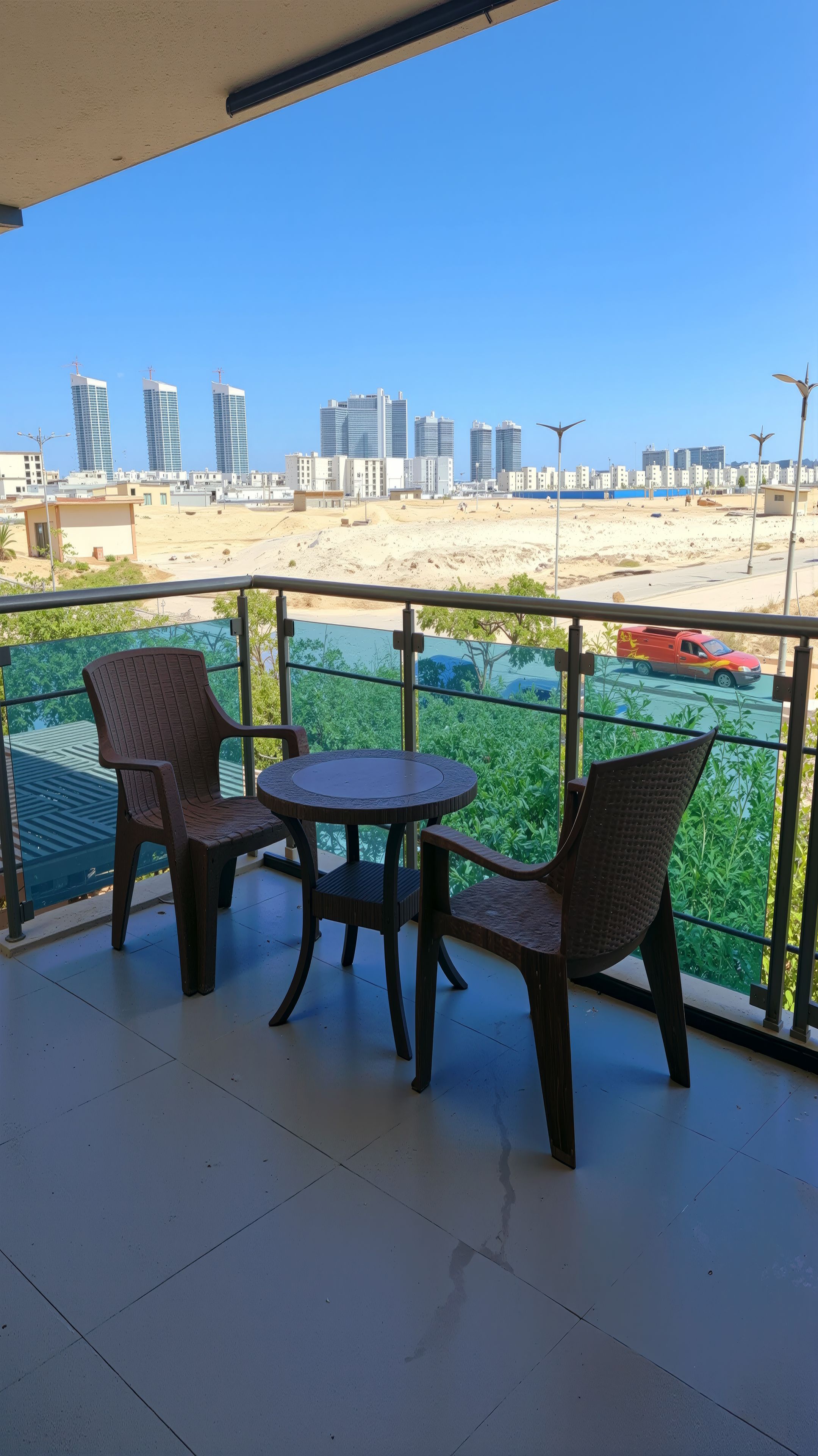 Un balcone con due sedie in vimini e un tavolino rotondo, affacciato su una spiaggia e su un paesaggio urbano con edifici moderni.